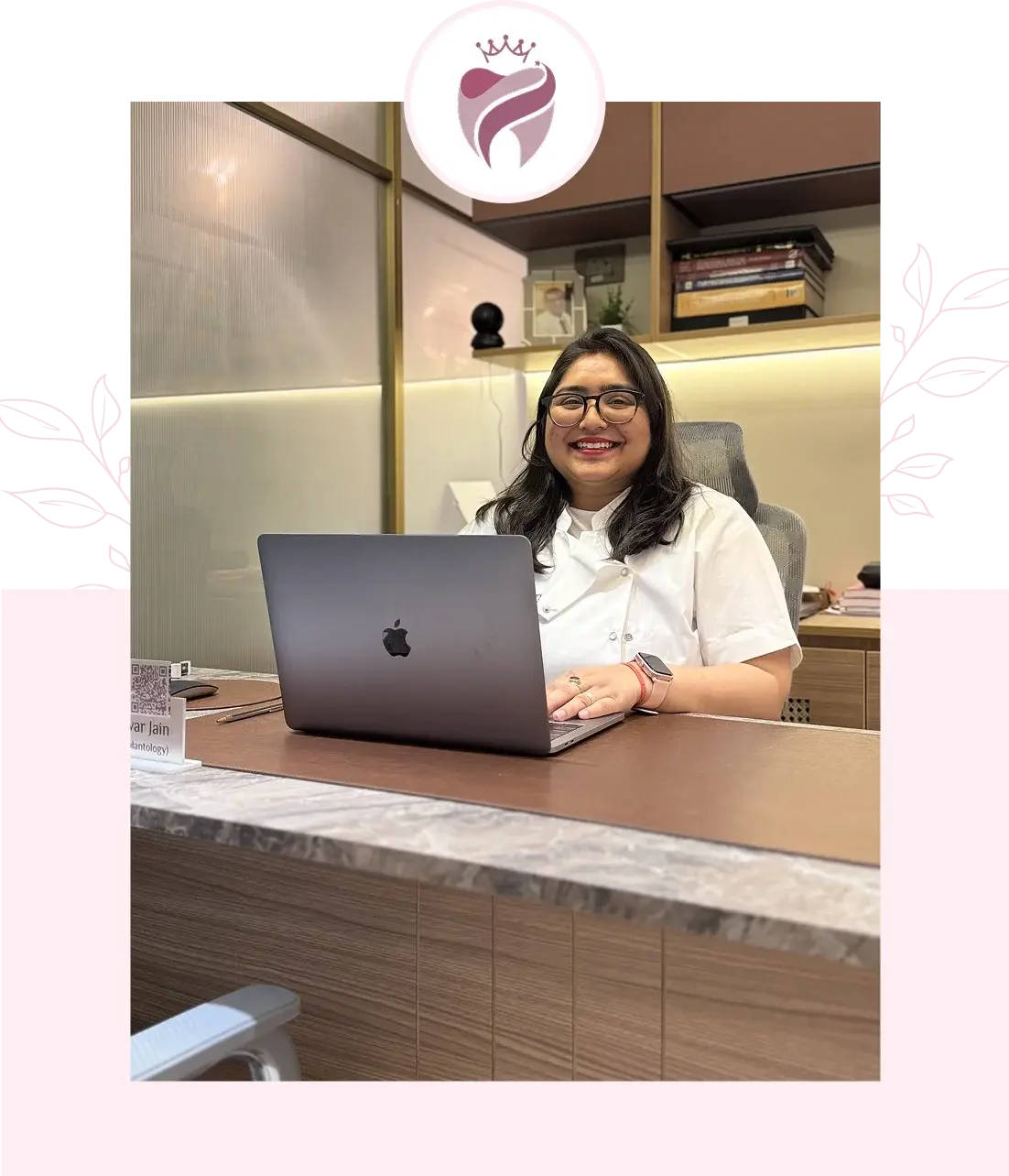 Dr. Jain smiling at her desk in a white coat, working on a MacBook in a modern dental clinic