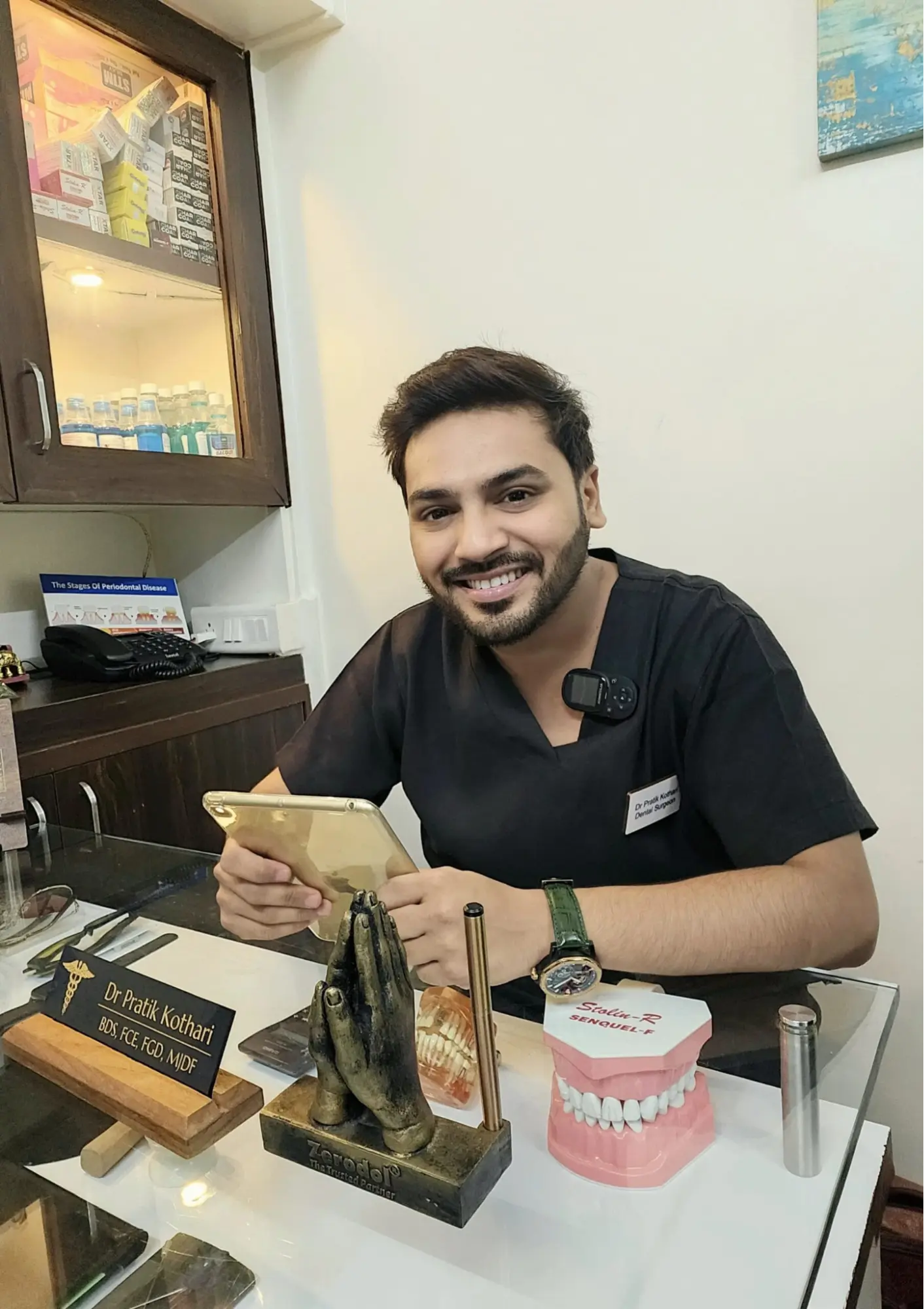Dr. Pratik Kothari seated at his clinic desk, holding a tablet, with dental models and a nameplate in view