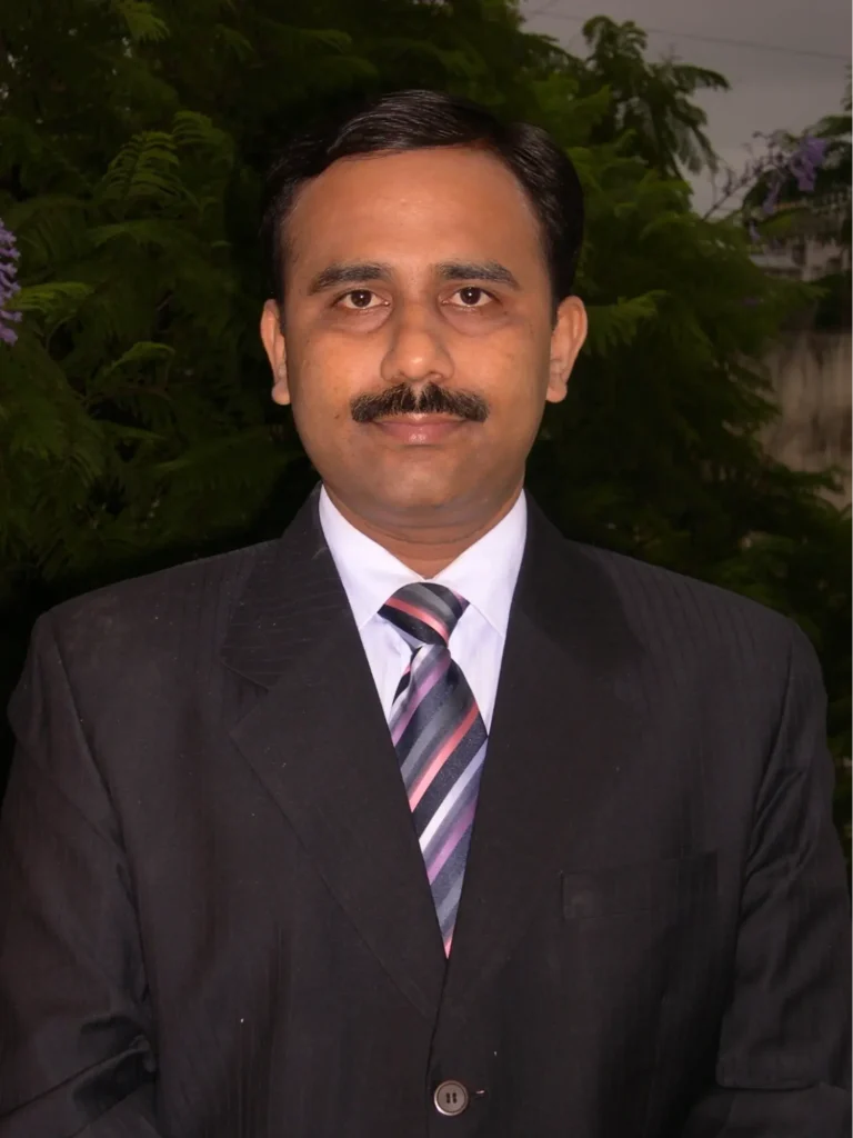 Professional headshot of a man in a formal black suit with a striped tie, standing in front of green foliage