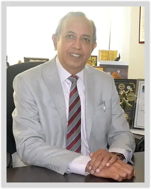 Portrait of a senior doctor seated at a desk in a grey suit with certificates and awards in the background